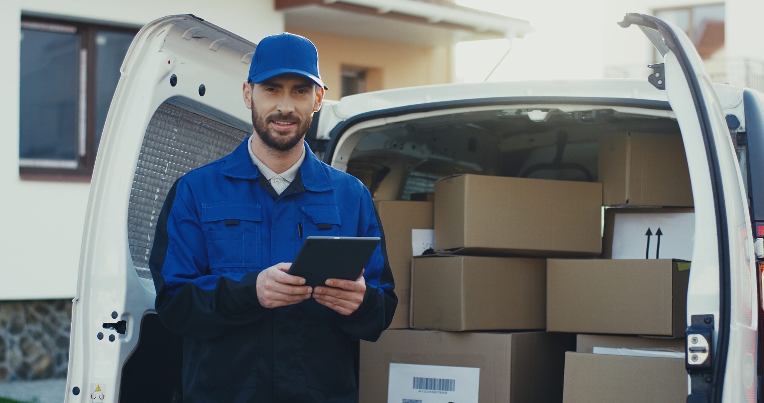 Portrait shot of the caucasian attractive deliveryman standing at the van full of parcels with a tablet computer in hands. Outside. Image of a delivery driver with a vehicle full of packages for peak delivery season
